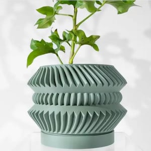 Può includere: Un vaso in ceramica verde salvia con un design geometrico. Il vaso ha una base larga e una parte superiore stretta, con una serie di creste e scanalature che creano una superficie testurizzata.