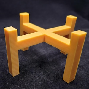 May include: A yellow-orange plant stand with a cross-shaped top and four vertical legs. The stand is made of a solid material and is designed to hold a potted plant. The stand is approximately 6 inches tall.