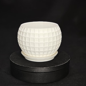 Può includere: Vaso da fiori in ceramica bianca con un motivo a griglia quadrata. Il vaso è rotondo e ha un sottovaso.