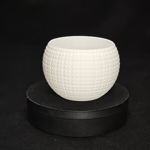 Può includere: Vaso in ceramica bianca con un motivo geometrico testurizzato. Il vaso è rotondo e si trova su un piedistallo nero.