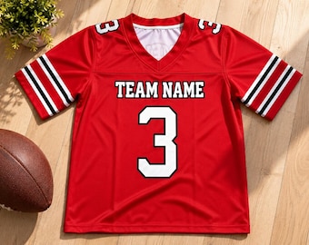 May include: A red V-neck sports jersey featuring the text TEAM NAME in black and white lettering, with the number 3 in white on the front and shoulders. The short sleeves have black and white stripes, and the fabric has a smooth, athletic finish.