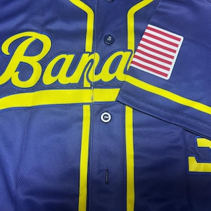 May include: A dark blue baseball jersey featuring the yellow script text "Bananas" across the chest. The jersey has yellow piping, dark blue buttons, and a patch of the United States flag on the right sleeve. A partial yellow number is visible below.