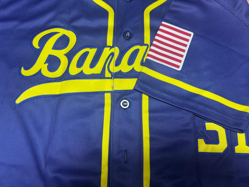 May include: A dark blue baseball jersey featuring the yellow script text "Bananas" across the chest. The jersey has yellow piping, dark blue buttons, and a patch of the United States flag on the right sleeve. A partial yellow number is visible below.