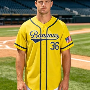 May include: A yellow baseball jersey with dark blue trim, featuring the word Bananas in script font across the chest, the number 36 below it, and a small American flag patch on the left sleeve. The button-down shirt has a rounded hemline.