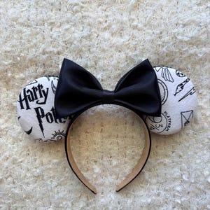 Puede incluir: Una diadema con dos orejas redondas blancas con ilustraciones de líneas negras que incluyen el texto Harry Potter y Platform 9 3/4, centrada con un gran lazo de satén negro sólido. La estructura de la diadema es de color beige neutro.