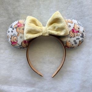 May include: A pair of mouse ears with a Winnie the Pooh theme. The ears are round and feature a patterned fabric with Winnie the Pooh illustrations. A large, fluffy yellow bow sits atop the headband. The headband is brown.