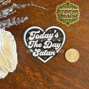 May include: A heart-shaped sticker with a black background and white border, featuring the white text Today's The Day Satan with small star accents. A US quarter coin is placed next to the sticker for scale, showing its relative size.