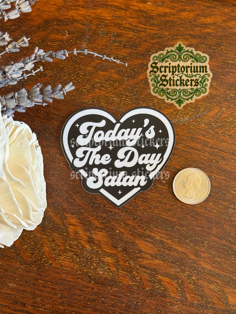 May include: A heart-shaped sticker with a black background and white border, featuring the white text Today's The Day Satan with small star accents. A US quarter coin is placed next to the sticker for scale, showing its relative size.