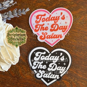 May include: Two heart-shaped stickers featuring the text Today's The Day Satan in a retro font. One sticker has a white background with a pink border and red text, while the other has a black background with a white border and white text.