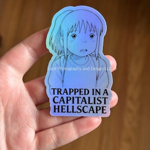 May include: Holographic sticker featuring an anime-style illustration of a girl with a surprised expression. The sticker has a blue and purple gradient and the text "TRAPPED IN A CAPITALIST HELLSCAPE" in white.