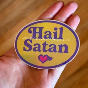 Pode incluir: Adesivo oval com as palavras "Hail Satan" em escrita roxa sobre um fundo amarelo. Uma borda roxa envolve o texto e um cora&ccedil;&atilde;o rosa est&aacute; abaixo da palavra "Satan". O adesivo &eacute; segurado em uma m&atilde;o.
