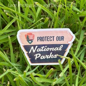 May include: A pink and brown sticker with the text "PROTECT OUR NATIONAL PARKS" and a National Park Service logo. The sticker is on a bed of green grass.