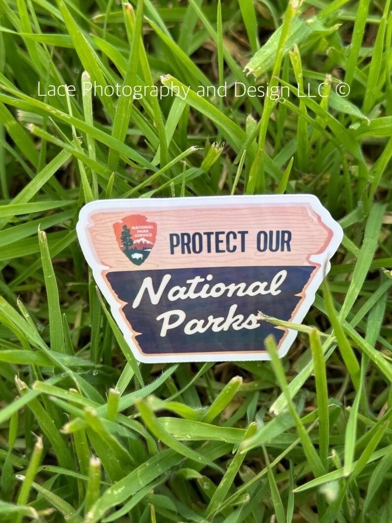 May include: A pink and brown sticker with the text "PROTECT OUR NATIONAL PARKS" and a National Park Service logo. The sticker is on a bed of green grass.