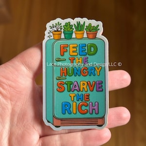 May include: A sticker featuring a teal refrigerator with the words "FEED THE HUNGRY STARVE THE RICH" in colorful text. The top of the refrigerator has four small potted plants. The sticker is held in a person's hand.