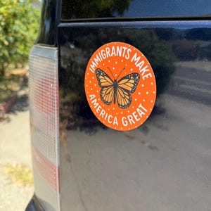 Puede incluir: Una pegatina redonda naranja con el texto "IMMIGRANTS MAKE AMERICA GREAT" y una ilustraci&oacute;n de una mariposa monarca. La pegatina est&aacute; adherida a la parte trasera de un veh&iacute;culo azul oscuro.