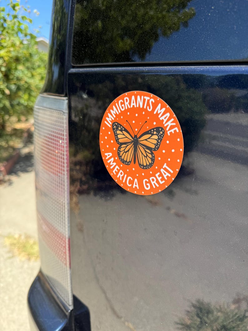 Puede incluir: Una pegatina redonda naranja con el texto "IMMIGRANTS MAKE AMERICA GREAT" y una ilustraci&oacute;n de una mariposa monarca. La pegatina est&aacute; adherida a la parte trasera de un veh&iacute;culo azul oscuro.