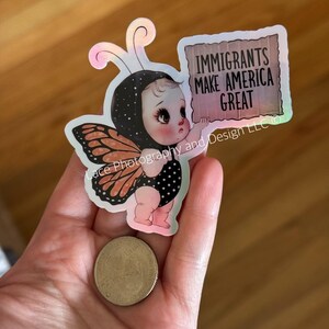 Puede incluir: Una pegatina que muestra a un beb&eacute; disfrazado de mariposa, sosteniendo un letrero que dice "IMMIGRANTS MAKE AMERICA GREAT". El beb&eacute; lleva un traje de lunares blancos y negros y alas de mariposa. La pegatina tiene un efecto hologr&aacute;fico.