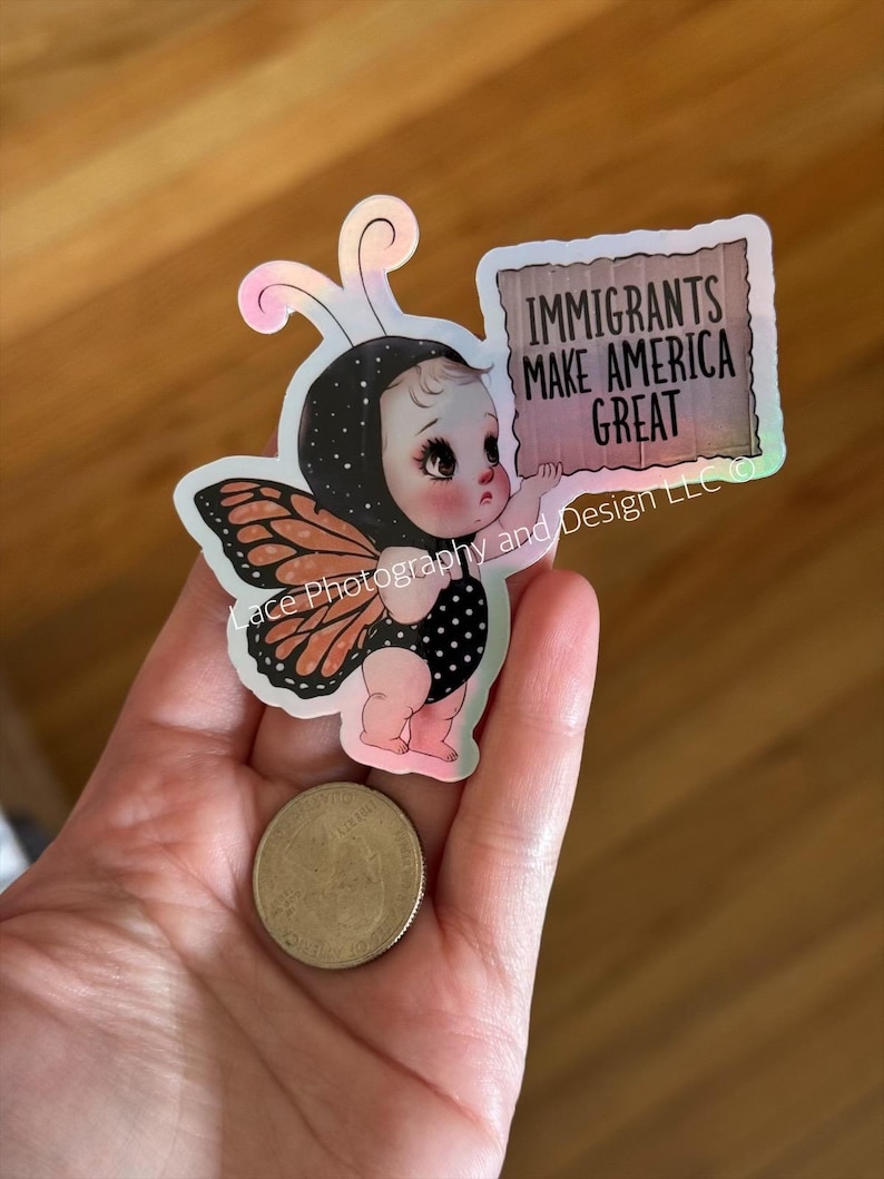 Puede incluir: Una pegatina que muestra a un beb&eacute; disfrazado de mariposa, sosteniendo un letrero que dice "IMMIGRANTS MAKE AMERICA GREAT". El beb&eacute; lleva un traje de lunares blancos y negros y alas de mariposa. La pegatina tiene un efecto hologr&aacute;fico.