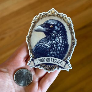 May include: A sticker with a detailed illustration of a raven in a gold frame. The raven is rendered in black and blue hues. A banner below the raven reads "I POOP ON FASCISTS".