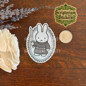 May include: A die-cut sticker featuring a black and white line art illustration of a rabbit wearing chainmail armor and holding a sword, enclosed in an ornate oval frame. A US quarter coin is placed next to the sticker for size reference.