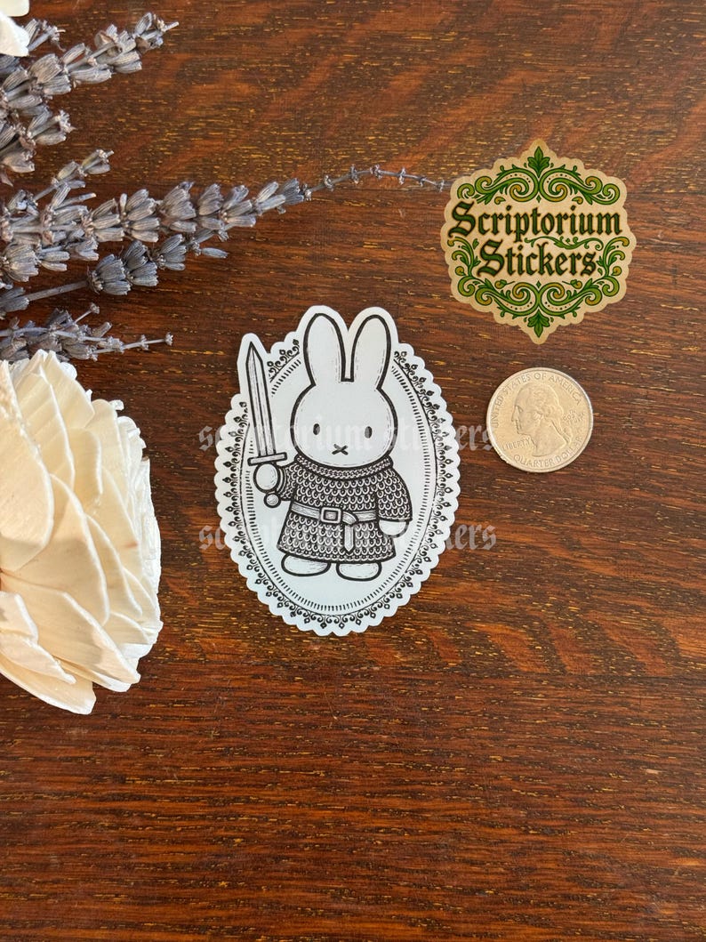 May include: A die-cut sticker featuring a black and white line art illustration of a rabbit wearing chainmail armor and holding a sword, enclosed in an ornate oval frame. A US quarter coin is placed next to the sticker for size reference.