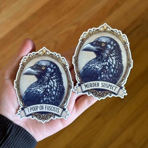 May include: Two stickers depicting a detailed illustration of a black raven in a gold frame. One reads "I POOP ON FASCISTS" and the other says "MURDER SUSPECT" on a banner. The stickers are held in a hand.