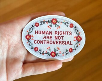 May include: Oval sticker with the text "HUMAN RIGHTS ARE NOT CONTROVERSIAL" in red letters. The text is surrounded by a floral wreath with red flowers and green leaves. The sticker is white with a white border.