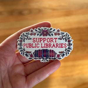 May include: An oval sticker with the text "SUPPORT PUBLIC LIBRARIES" in red pixelated letters. The sticker is framed by a floral border with red and pink flowers and green leaves. A row of colorful books is below the text.