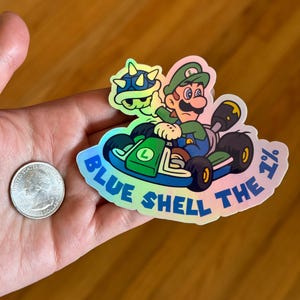 May include: A holographic sticker featuring a green cartoon character driving a kart with a blue shell. The text "Blue Shell the 1%" is written around the sticker.