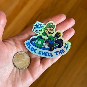 May include: A holographic sticker featuring a green-clad Mario character driving a kart with a blue shell. The sticker reads "Blue Shell the 1%".
