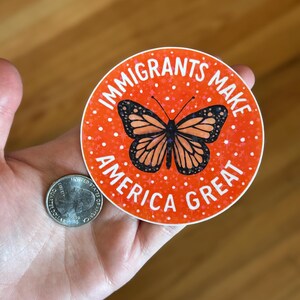 Puede incluir: Una pegatina redonda con el texto "IMMIGRANTS MAKE AMERICA GREAT" en blanco sobre un fondo naranja. Un gr&aacute;fico de mariposa monarca est&aacute; en el centro. Una moneda estadounidense est&aacute; al lado de la pegatina.