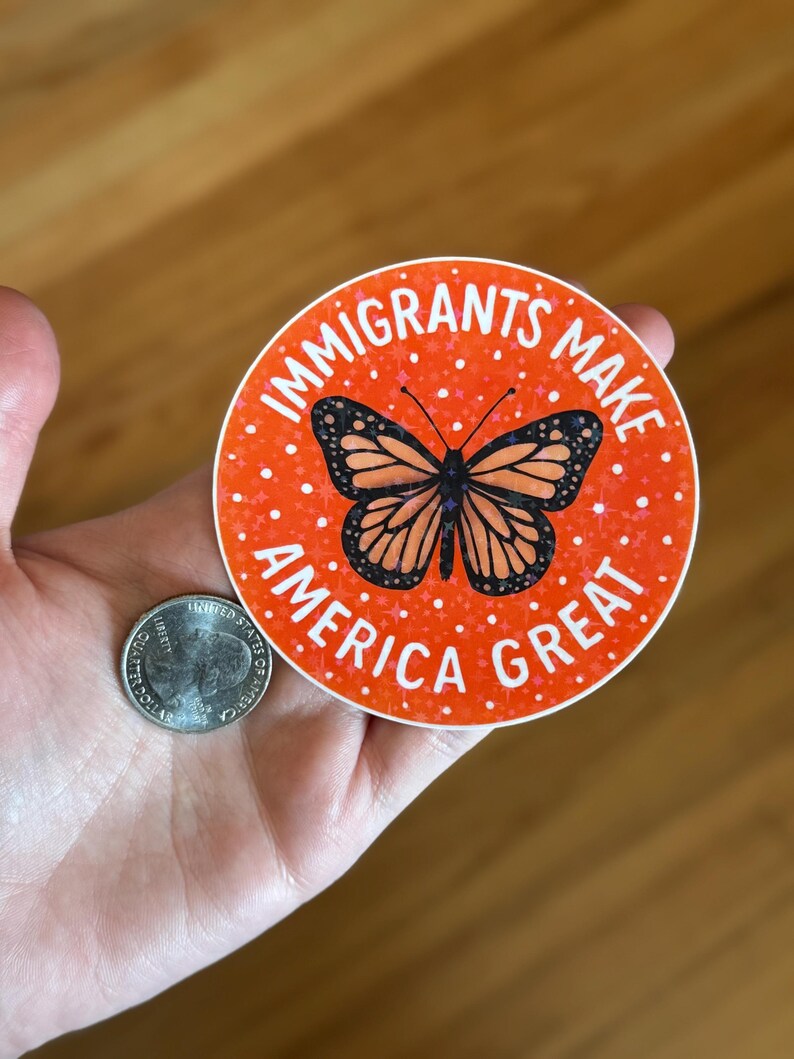 Puede incluir: Una pegatina redonda con el texto "IMMIGRANTS MAKE AMERICA GREAT" en blanco sobre un fondo naranja. Un gr&aacute;fico de mariposa monarca est&aacute; en el centro. Una moneda estadounidense est&aacute; al lado de la pegatina.
