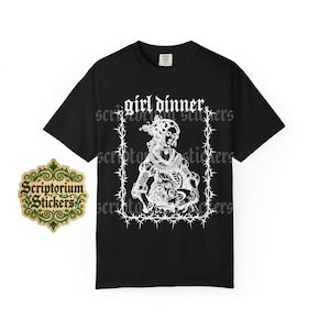 May include: A black short-sleeve t-shirt featuring a white graphic print of a historical figure within a barbed wire frame. The text "girl dinner" is printed in a gothic-style font above the illustration. The shirt has a crew neckline.