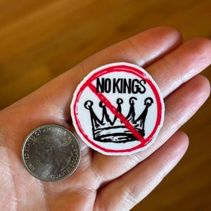 May include: A round sticker with the text "NO KINGS" in red and black, with a crown design crossed out. A United States quarter coin is next to the sticker.