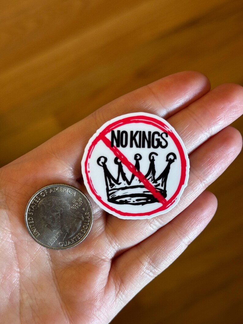 May include: A round sticker with the text "NO KINGS" in red and black, with a crown design crossed out. A United States quarter coin is next to the sticker.