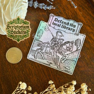 May include: A sticker featuring the text "Defend thy local library" above a black and white illustration of a knight on horseback. A green and gold sticker with "Scriptorium Stickers" is also visible.
