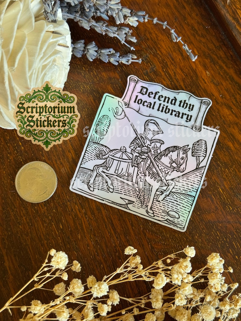 May include: A sticker featuring the text "Defend thy local library" above a black and white illustration of a knight on horseback. A green and gold sticker with "Scriptorium Stickers" is also visible.