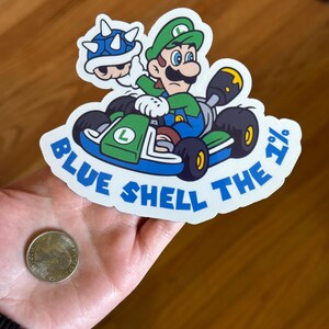 May include: A white sticker with a blue shell, green kart, and a green Mario character driving. The text on the sticker reads "Blue Shell The 1%".