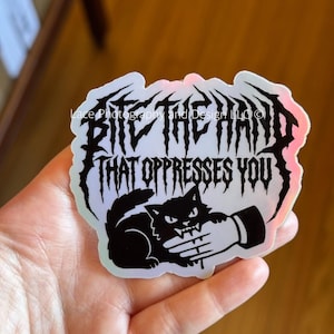 May include: A holographic sticker with the text "BITE THE HAND THAT OPPRESSES YOU" in a gothic font. The sticker features a black cat biting a hand. The sticker has a white border and a holographic sheen.