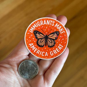 Puede incluir: Una pegatina redonda con el texto "IMMIGRANTS MAKE AMERICA GREAT" en blanco sobre un fondo naranja. Un gr&aacute;fico de mariposa monarca en el centro. Una moneda de veinticinco centavos estadounidense est&aacute; al lado.