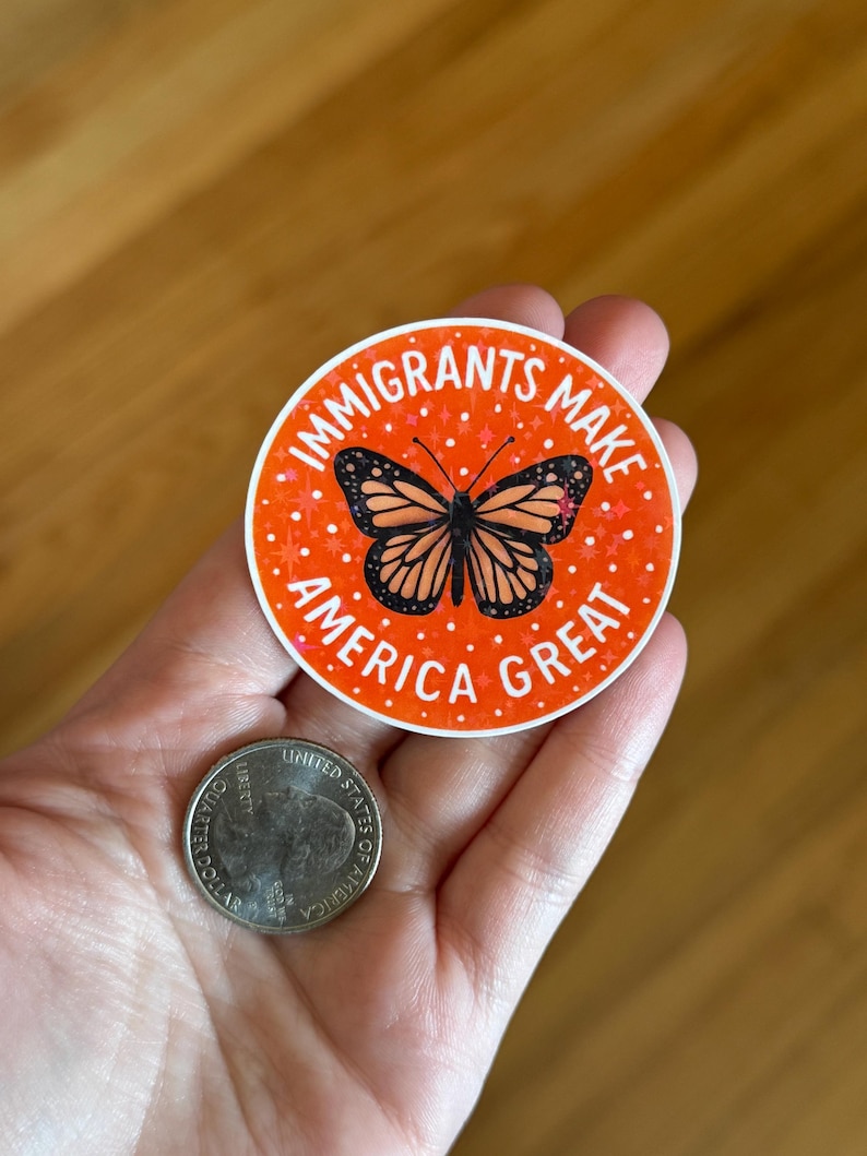 Puede incluir: Una pegatina redonda con el texto "IMMIGRANTS MAKE AMERICA GREAT" en blanco sobre un fondo naranja. Un gr&aacute;fico de mariposa monarca en el centro. Una moneda de veinticinco centavos estadounidense est&aacute; al lado.