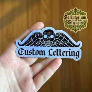 May include: A holographic sticker with a skull and wing design, and the words "Custom Lettering". The sticker has a black and white color scheme with a light blue gradient. A second sticker with the text "Scriptorium Stickers" is also visible.