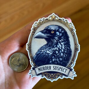 May include: A sticker with a detailed illustration of a raven's head within an ornate, gold-framed oval. The raven is depicted in shades of blue and black with a yellow eye. Below the frame is a banner that reads "MURDER SUSPECT".