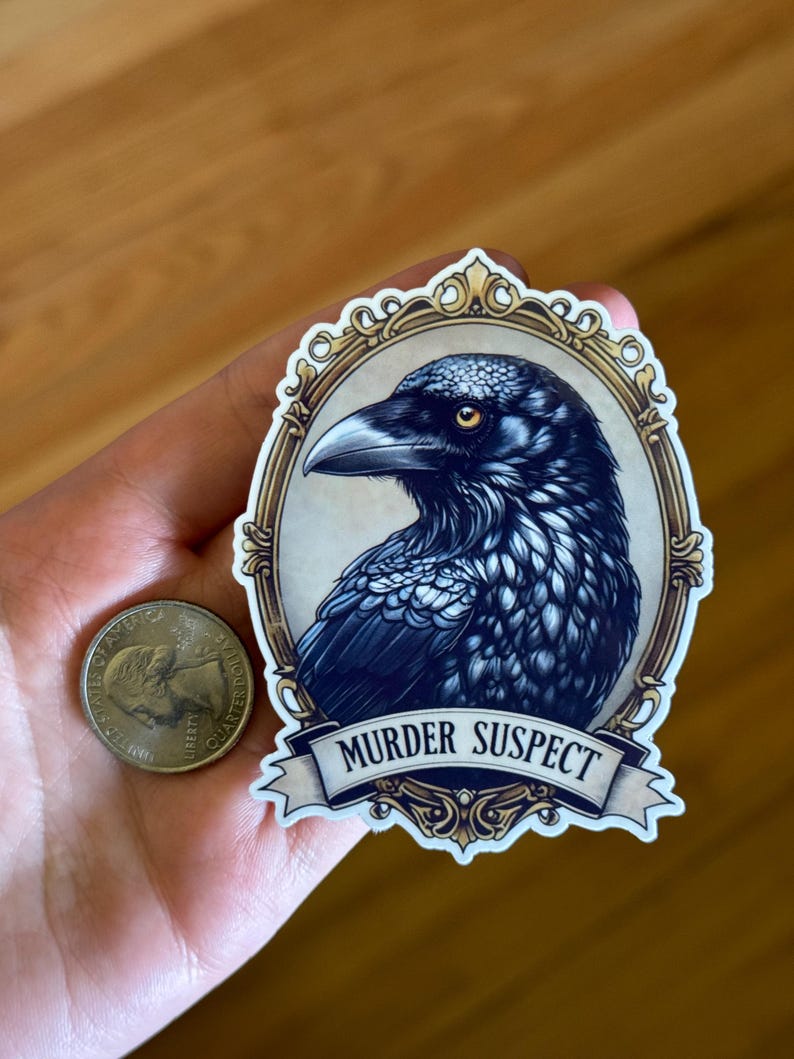 May include: A sticker with a detailed illustration of a raven's head within an ornate, gold-framed oval. The raven is depicted in shades of blue and black with a yellow eye. Below the frame is a banner that reads "MURDER SUSPECT".