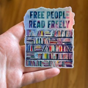 May include: A sticker with the text "FREE PEOPLE READ FREELY" above a watercolor illustration of a bookshelf filled with colorful books. The sticker has a watercolor effect with blue, purple, and pink hues.