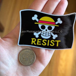 May include: A black sticker with a white skull and crossbones design wearing a yellow and red hat, with the word "RESIST" in yellow. A U.S. quarter coin is next to the sticker.