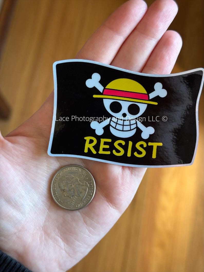 May include: A black sticker with a white skull and crossbones design wearing a yellow and red hat, with the word "RESIST" in yellow. A U.S. quarter coin is next to the sticker.