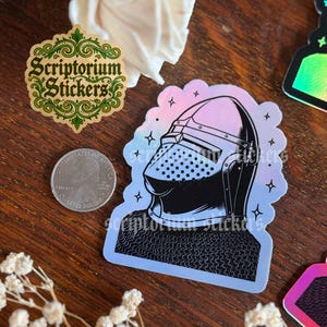 May include: A collection of stickers on a wooden surface. One sticker features a medieval knight's helmet in black and white with a holographic effect. Another sticker has the text "Scriptorium Stickers" in green and gold. A U.S. quarter is also visible.