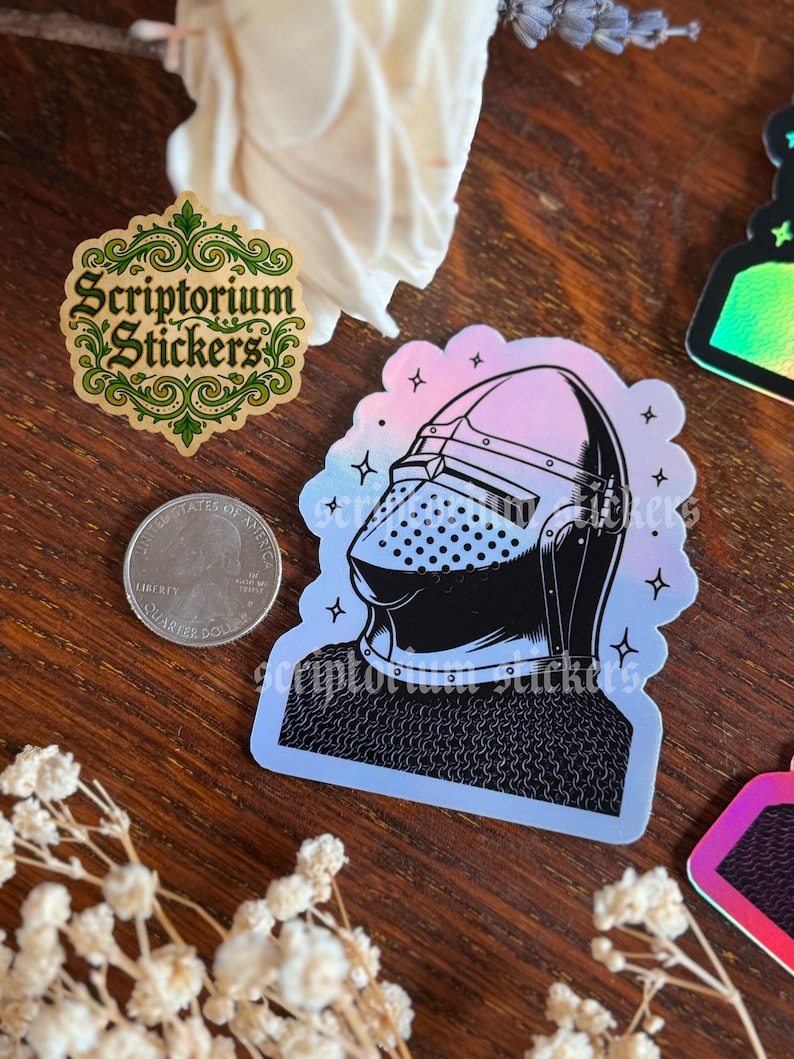 May include: A collection of stickers on a wooden surface. One sticker features a medieval knight's helmet in black and white with a holographic effect. Another sticker has the text "Scriptorium Stickers" in green and gold. A U.S. quarter is also visible.