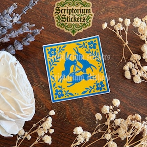 May include: A square sticker with a yellow background and blue floral and rabbit design. The sticker has a blue border and the text "Scriptorium Stickers" in a green and gold design.